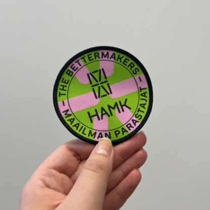 Pyöreä merkki, jossa väreinä vaaleanpunainen ja vihreä. Keskellä musta HAMK logo ja kehällä mustalla tekstit "The bettermakers" ja "Maailman parastajat"